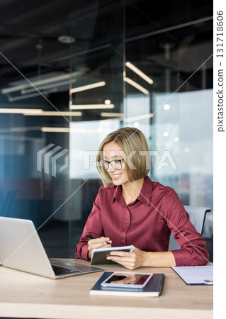 Smiling businesswoman in glasses taking notes while working on a laptop at her desk in a contemporary office, representing productivity and professional success 131718606