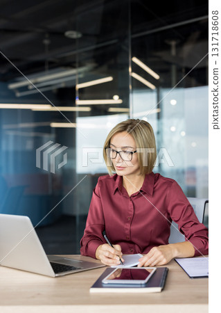 Businesswoman wearing glasses works at a laptop in a modern corporate office, taking notes in a notebook while concentrating on planning, strategy and project tasks at her desk Businesswoman wearing glasses works at a laptop in a modern corporate office, taking notes in a notebook while concentrating on planning, strategy and project tasks at her desk 131718608