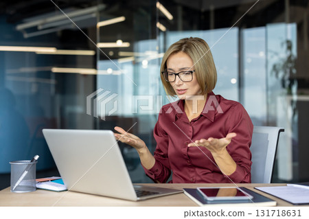 Businesswoman gesturing while having a video call on a laptop in a modern office, expressing concern and confusion during an online meeting 131718631