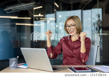 Enthusiastic businesswoman is raising her fists in celebration while sitting at her desk in a modern office, expressing joy and excitement over a successful achievement on her laptop Enthusiastic businesswoman is raising her fists in celebration while sitting at her desk in a modern office, expressing joy and excitement over a successful achievement on her laptop 131718634