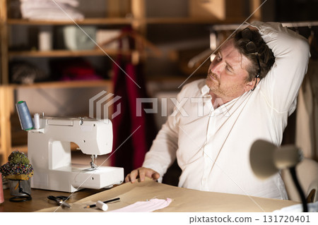 Middle-aged male tailor stretching while sitting next to sewing machine at night Middle-aged male tailor stretching while sitting next to sewing machine at night 131720401
