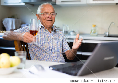 Elderly man with glass of wine communicates virtually using a laptop and the Internet 131720412