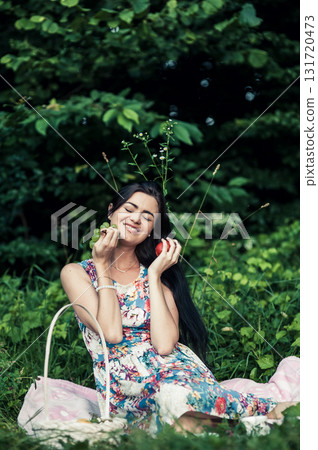 A young girl is sitting on a pink rug, next to a white basket with green grapes, nectarines, and apples 131720473