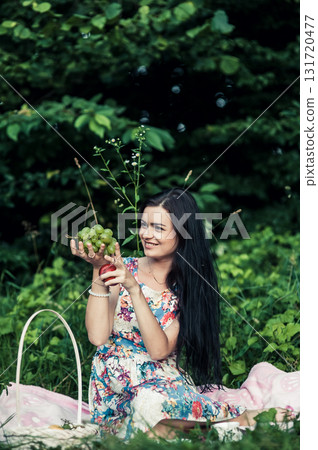 A woman in a bright floral dress sits on a pink rug during a picnic A woman in a bright floral dress sits on a pink rug during a picnic 131720477