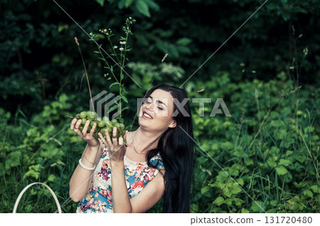 A young girl at a picnic in the park holds a large bunch of grapes against a backdrop of green trees 131720480