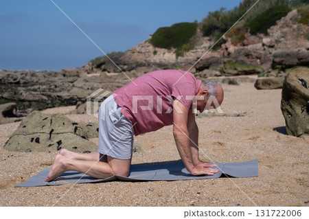 Elderly man practicing yoga on the beach doing cat pose for back health 131722006