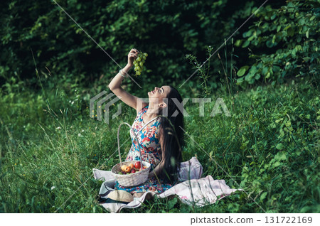 A lovely woman sits on a pink rug during a picnic, with a white basket of fruit standing nearby A lovely woman sits on a pink rug during a picnic, with a white basket of fruit standing nearby 131722169
