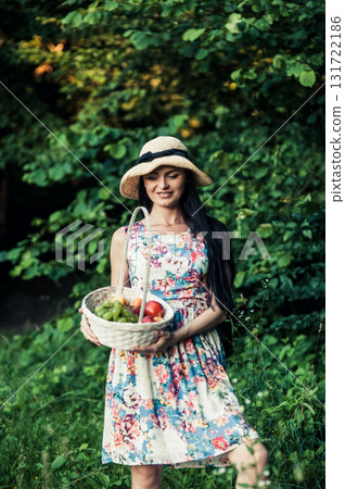A lady in a floral dress with a cute hat is standing at a picnic next to a pink rug 131722186
