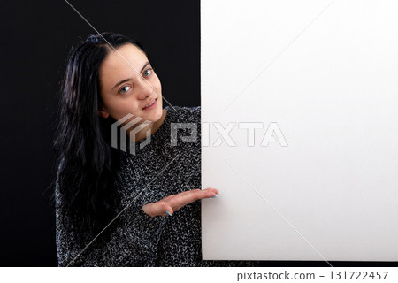 attractive dark-haired woman in a gray sweater with a white blank poster, isolated on a dark 131722457