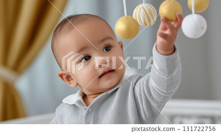 Baby reaching for hanging mobile decorations with curiosity and delight. Indoor nursery setting creates a warm, inviting atmosphere. Concept of childcare, early development, parenting. 131722756