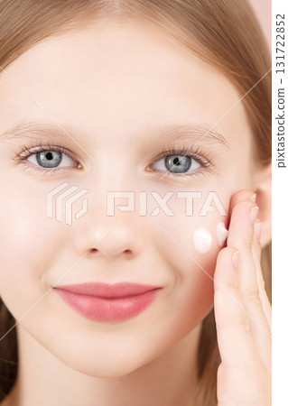 Close-up portrait of a smiling cheerful girl applying face cream on cheek for skin glow. Gen Alpha. Ad face cream and skincare products for teens. Self care. Blonde girl with blue eyes. Studio shot 131722852
