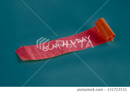 A red textured paper with white handwriting that says SLAY ALL DAY is partially unrolled against a Teal background in a studio setting, promoting confidence and goals 131723531