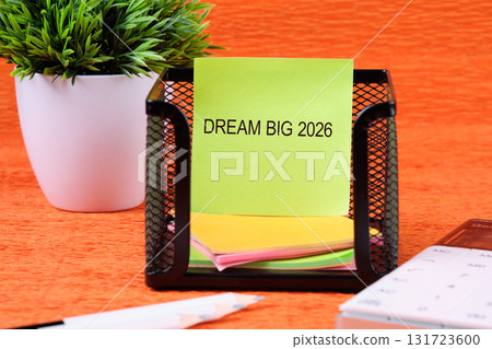 A light green note reading DREAM BIG 2026 sits in a black holder next to a plant, pencil, and calculator on an orange desk. It seems like a business related setting 131723600