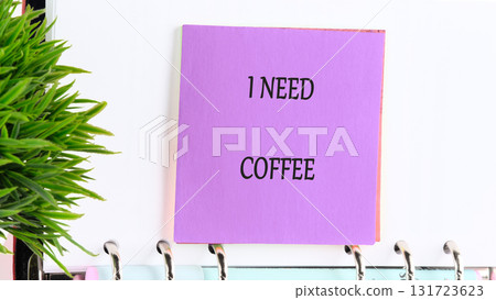 A purple sticky note with the words I NEED COFFEE is attached to a spiral notebook with a green artificial plant situated next to it, craving its morning or afternoon pick-me-up A purple sticky note with the words I NEED COFFEE is attached to a spiral notebook with a green artificial plant situated next to it, craving its morning or afternoon pick-me-up 131723623