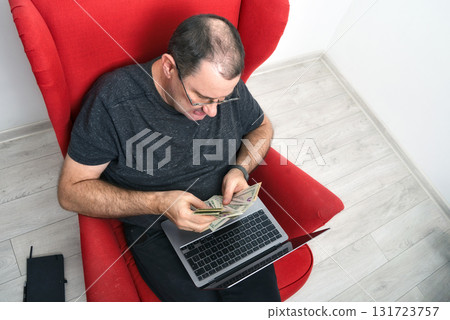 middle-aged man in glasses with a laptop on his lap sitting in a red chair and counting money middle-aged man in glasses with a laptop on his lap sitting in a red chair and counting money 131723757