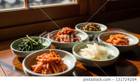 Six traditional Korean banchan side dishes arranged in ceramic bowls on wooden table by window featuring kimchi pickled vegetables spinach and radish 131724204