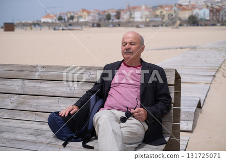 Elderly man sitting on a bench at the beach, resting and sunbathing 131725071