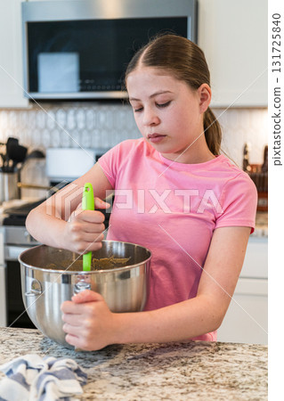 A girl in a pink shirt focuses on mixing colorful cookie dough in a large metal bowl. The background shows a white kitchen with cabinets and countertop appliances. A girl in a pink shirt focuses on mixing colorful cookie dough in a large metal bowl. The background shows a white kitchen with cabinets and countertop appliances. 131725840