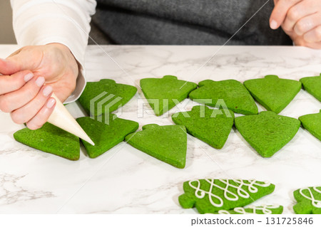 A workspace featuring rows of green Christmas tree cookies, ready for decorating, with decorating tools visible in the background. A workspace featuring rows of green Christmas tree cookies, ready for decorating, with decorating tools visible in the background. 131725846
