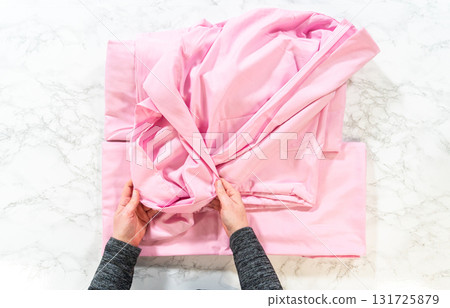 Close-up top-down view of hands folding a soft pink bedsheet on a marble surface. The image captures the technique of tucking edges neatly. Close-up top-down view of hands folding a soft pink bedsheet on a marble surface. The image captures the technique of tucking edges neatly. 131725879
