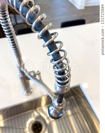 A close-up image of a modern industrial-style kitchen faucet with a flexible coil neck over a stainless steel sink. The faucet is installed on a white quartz countertop in a contemporary kitchen 131725884