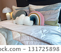 A cozy bed is decorated with rainbow and cloud shaped pillows. The room features modern lamps and a comfortable, cheerful interior design style. 131725898