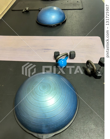 A workout station featuring a bosu ball, kettlebell, dumbbells, and a mat set up indoors. The equipment is placed in an orderly manner under bright lighting, creating a clean and structured workout 131725907
