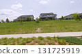 A paved sidewalk runs beside a mowed green field in a residential area. Houses with decks and fencing are visible across the space. 131725919