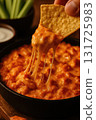 A tortilla chip lifts a serving of hot melted cheese dip from a skillet. The cheese stretches creating a gooey texture. The close up shot emphasizes indulgence and warmth in a casual food setting. 131725983