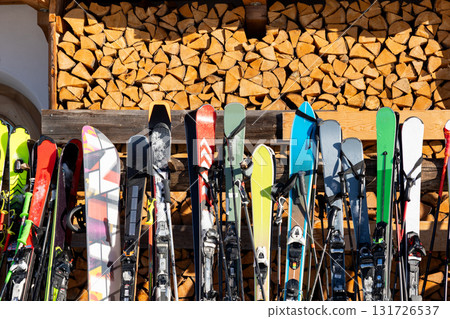 Colorful skis snowboards stand row against rustic wooden wall stacked with chopped firewood. Bright sunlight highlights vivid equipment colors warm wood tones, creating cozy alpine winter atmosphere 131726537