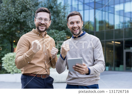 Two businessmen celebrate outdoors in front of a modern office building, cheering with raised fists and a tablet, smiling confidently after a successful deal and teamwork triumph 131726716