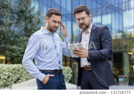 Two stressed businessmen stand outside an office building, studying a tablet with concerned, puzzled expressions as they analyze data and discuss a difficult corporate problem 131726775