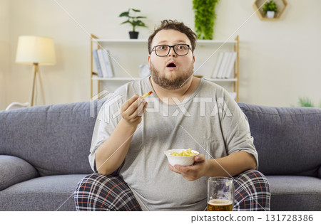 Overweight man eating fries on sofa, watching TV with beer at home, late evening 131728386
