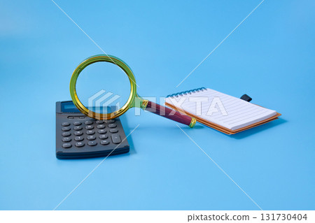 Magnifier glass with calculator and a notebook on background. Magnifier glass with calculator and a notebook on background. 131730404