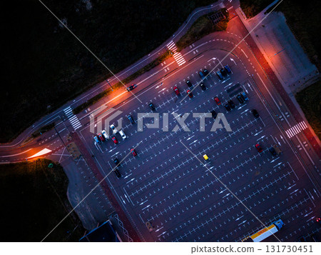 Aerial twilight view of organized parking lot warm streetlights illuminate rows of vehicles, crosswalks, and pathways leading into grassy terrain, blending urban geometry with natural contours. 131730451