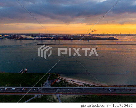 Aerial sunset view of coastal industrial zone road, beach, and grassy shoreline foreground contrast with smokestacks, cranes, and breakwater across golden water under dramatic cloudscape. 131730455