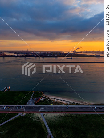 Aerial sunset view of coastal industrial zone road, beach, and grassy shoreline foreground contrast with smokestacks, cranes, and breakwater across golden water under dramatic cloudscape. 131730456
