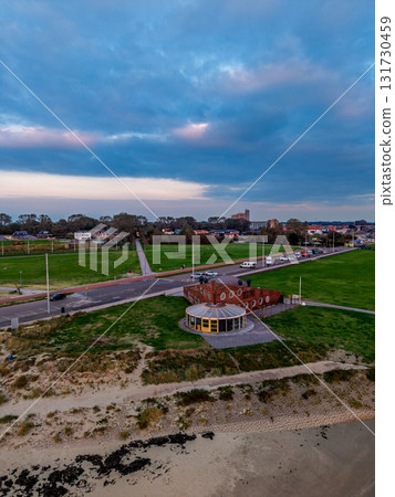 Aerial view of suburban zone at dusk circular brick building with yellow entrance anchors grassy roadside, while vehicles and dense urban blocks stretch beneath dramatic cloud filled sky. 131730459