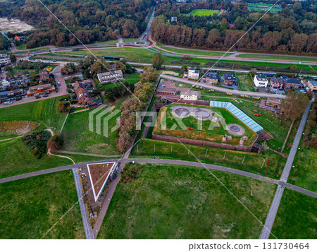 Aerial view of semi rural zone angular building with rooftop circles and green roofed triangle structure amid grassy fields, roads, forest edge, and sports field near clustered homes. Aerial view of semi rural zone angular building with rooftop circles and green roofed triangle structure amid grassy fields, roads, forest edge, and sports field near clustered homes. 131730464