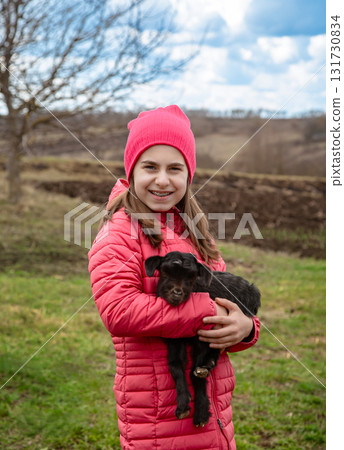 Smiling girl in red jacket and beanie holding black baby goat on farm. Rural lifestyle, childhood, animal care countryside setting Smiling girl in red jacket and beanie holding black baby goat on farm. Rural lifestyle, childhood, animal care countryside setting 131730834