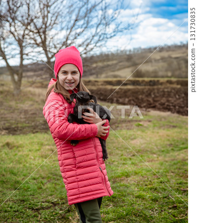 Smiling girl in red jacket and beanie holding black baby goat on farm. Rural lifestyle Smiling girl in red jacket and beanie holding black baby goat on farm. Rural lifestyle 131730835