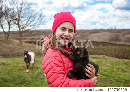 Smiling girl in red jacket and beanie holding black baby goat on farm. Rural lifestyle Smiling girl in red jacket and beanie holding black baby goat on farm. Rural lifestyle 131730839