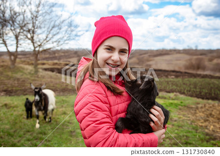 Smiling girl in red jacket and beanie holding black baby goat on farm. Rural lifestyle, childhood, animal care countryside setting 131730840