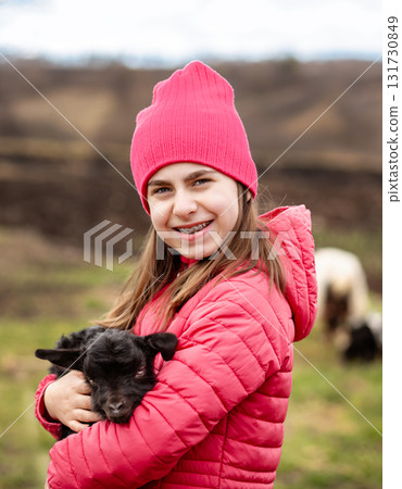 Cute girl in red jacket and beanie holding black baby goat on farm. Rural lifestyle, childhood, animal care countryside setting Cute girl in red jacket and beanie holding black baby goat on farm. Rural lifestyle, childhood, animal care countryside setting 131730849