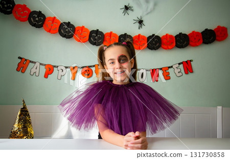 Smiling girl purple tulle costume with Halloween makeup, sitting decorated room with pumpkins, garlands, spiders, and witch hat. Smiling girl purple tulle costume with Halloween makeup, sitting decorated room with pumpkins, garlands, spiders, and witch hat. 131730858