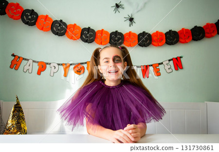 Smiling girl purple tulle costume with Halloween makeup, sitting decorated room with pumpkins, garlands, spiders, and witch hat. Smiling girl purple tulle costume with Halloween makeup, sitting decorated room with pumpkins, garlands, spiders, and witch hat. 131730861