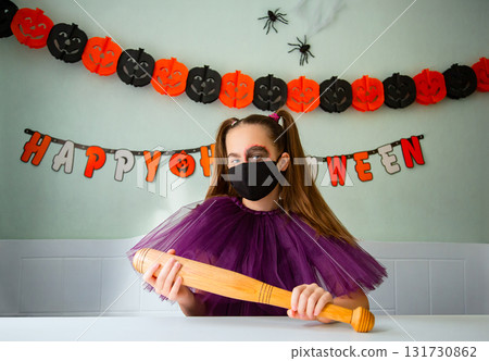 Girl dressed in purple costume with face makeup and black face mask holds wooden bat in room decorated for Halloween. 131730862
