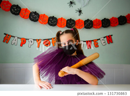 Girl dressed in purple costume with face makeup and black face mask holds wooden bat in room decorated for Halloween. Girl dressed in purple costume with face makeup and black face mask holds wooden bat in room decorated for Halloween. 131730863