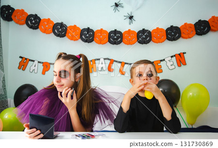 Two kids getting ready for Halloween. One girl applies makeup while boy inflates balloon. Two kids getting ready for Halloween. One girl applies makeup while boy inflates balloon. 131730869