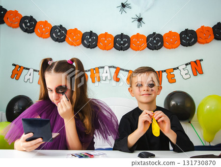 Two kids getting ready for Halloween. One girl applies makeup while boy inflates balloon. 131730870